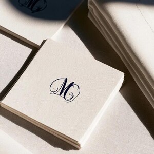 Square navy and cream cocktail napkins with small contrasting monogram letters, displayed with coordinating paper pieces. Digital concept mockup showing a personalised wedding or event table detail.