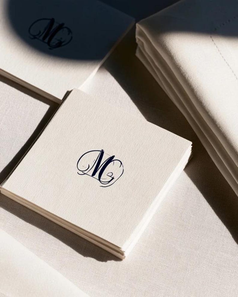 Square navy and cream cocktail napkins with small contrasting monogram letters, displayed with coordinating paper pieces. Digital concept mockup showing a personalised wedding or event table detail.