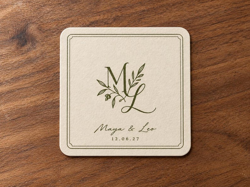Digital tabletop mockup of one thin ivory square paper coaster with olive-green ML monogram, Maya and Leo names and date, lying on a simple walnut table. Standard printed wedding design example.