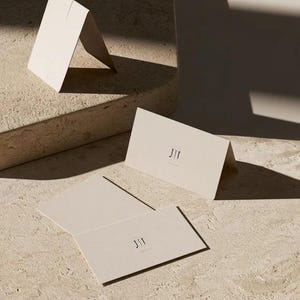 Cream wedding place cards with small dark initials and fine lettering, shown as flat and folded cards on a pale surface. Digital concept mockup of personalised table stationery.
