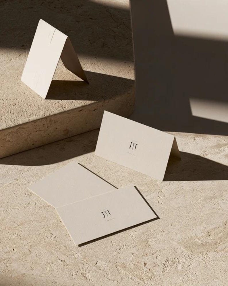 Cream wedding place cards with small dark initials and fine lettering, shown as flat and folded cards on a pale surface. Digital concept mockup of personalised table stationery.