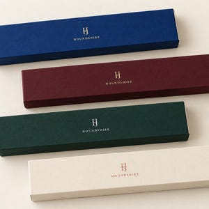 Digital colour and finish concept showing four closed extra-long Houndshire matchboxes: navy and burgundy with gold foil, forest green with silver foil, and ivory with copper foil, on a plain light surface.