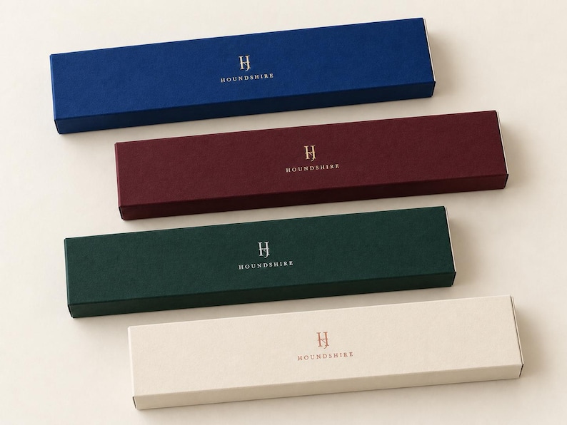 Digital colour and finish concept showing four closed extra-long Houndshire matchboxes: navy and burgundy with gold foil, forest green with silver foil, and ivory with copper foil, on a plain light surface.