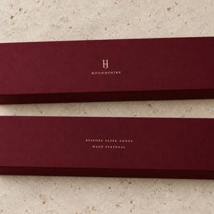 Digital design mockup of two closed burgundy extra-long matchboxes with gold-foil Houndshire lettering, showing front and reverse on pale travertine. Metallic foil is an optional upgrade.