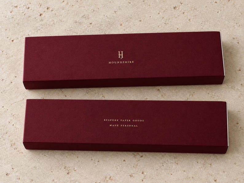 Digital design mockup of two closed burgundy extra-long matchboxes with gold-foil Houndshire lettering, showing front and reverse on pale travertine. Metallic foil is an optional upgrade.