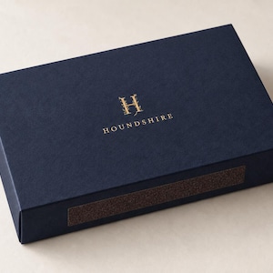Digital close-up mockup of a closed navy drawer matchbox with gold-foil Houndshire monogram, fine paper texture, folded edge and brown striker panel. Optional paid foil finish; not a physical sample photograph.