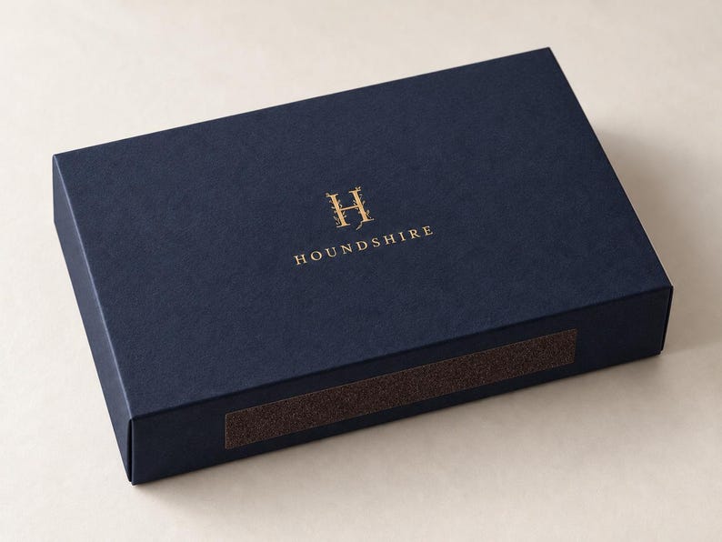 Digital close-up mockup of a closed navy drawer matchbox with gold-foil Houndshire monogram, fine paper texture, folded edge and brown striker panel. Optional paid foil finish; not a physical sample photograph.