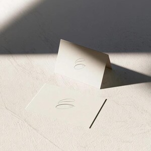 Cream wedding place cards with small dark initials and fine lettering, shown as flat and folded cards on a pale surface. Digital concept mockup of personalised table stationery.