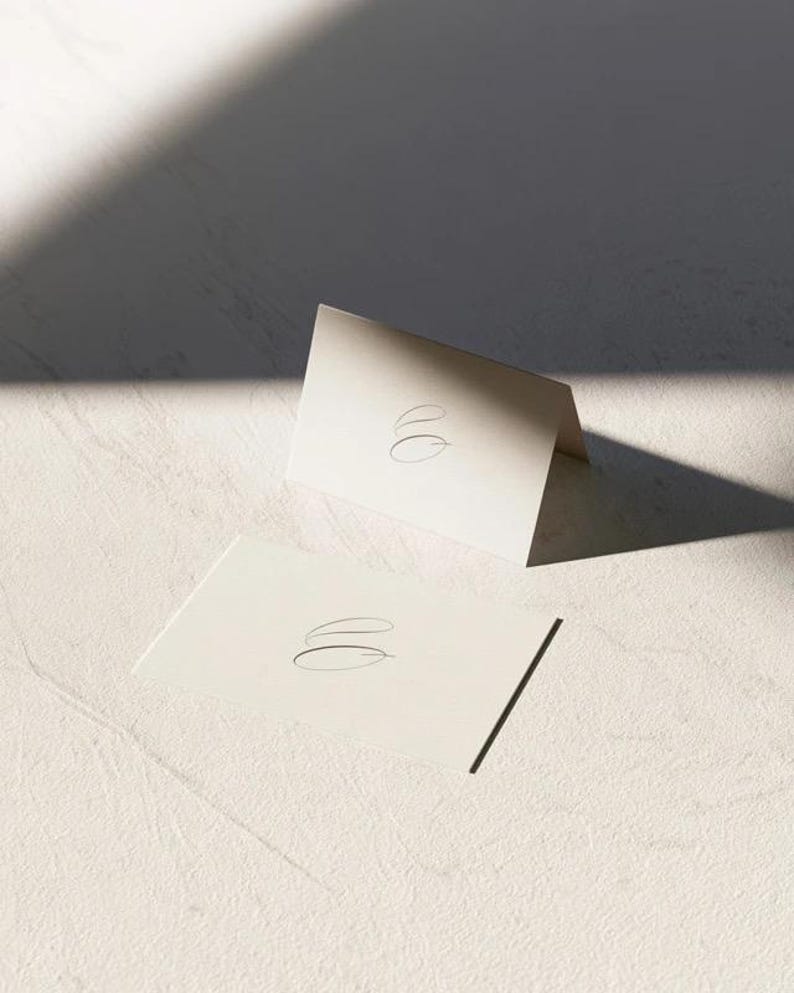 Cream wedding place cards with small dark initials and fine lettering, shown as flat and folded cards on a pale surface. Digital concept mockup of personalised table stationery.