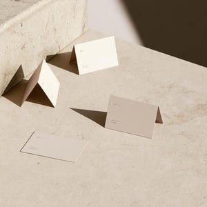 Cream wedding place cards with small dark initials and fine lettering, shown as flat and folded cards on a pale surface. Digital concept mockup of personalised table stationery.