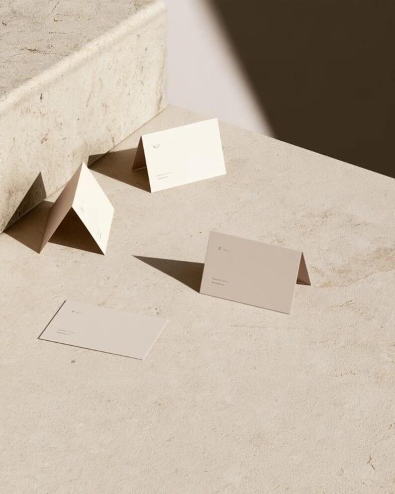 Cream wedding place cards with small dark initials and fine lettering, shown as flat and folded cards on a pale surface. Digital concept mockup of personalised table stationery.