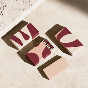 May include: Four rectangular matchboxes with a cream and deep red abstract pattern. One box is partially open, revealing a row of matches with dark red heads. The boxes are arranged in a cluster, with one plain cream-colored box lid placed separately.
