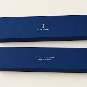 Digital design mockup of two closed navy extra-long matchboxes with a small gold-foil Houndshire monogram and reverse wording, photographed against a warm ivory studio background. Foil is an optional upgrade.