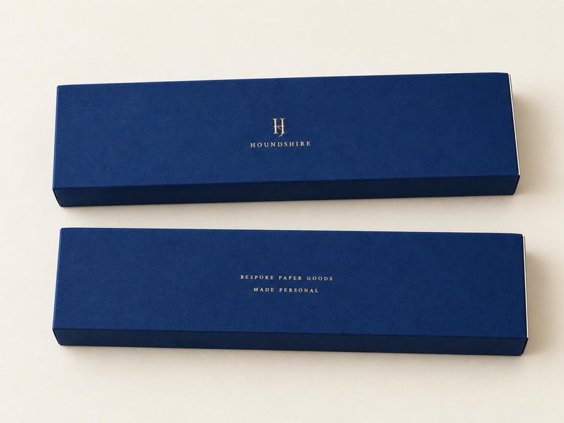 Digital design mockup of two closed navy extra-long matchboxes with a small gold-foil Houndshire monogram and reverse wording, photographed against a warm ivory studio background. Foil is an optional upgrade.