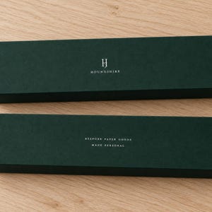 Digital design mockup of two closed forest-green extra-long matchboxes with silver-foil Houndshire lettering and reverse wording on a light oak tabletop. Foil is an optional upgrade; no matches visible.