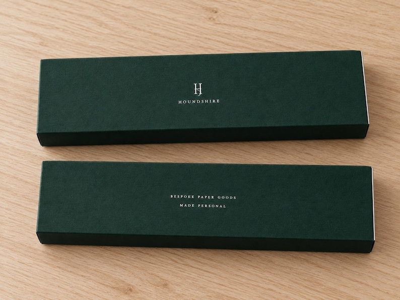 Digital design mockup of two closed forest-green extra-long matchboxes with silver-foil Houndshire lettering and reverse wording on a light oak tabletop. Foil is an optional upgrade; no matches visible.