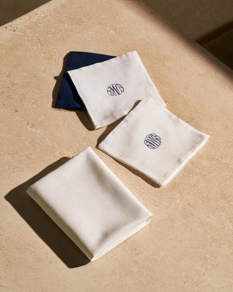 Square navy and cream cocktail napkins with small contrasting monogram letters, displayed with coordinating paper pieces. Digital concept mockup showing a personalised wedding or event table detail.