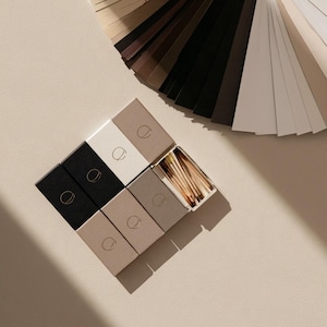 Small rectangular matchbox mockups in a restrained neutral palette, decorated with fine monograms. The styled arrangement shows the paper sleeves and drawer-style construction for personalised event favours.