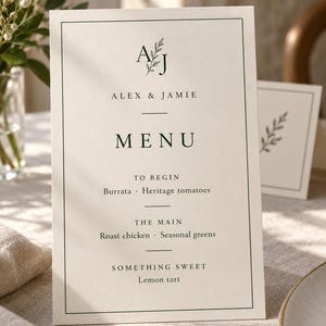 Ivory wedding menu card with a green AJ monogram, fine border, and a legible three-course sample menu. Styled on a linen table beside a plate. Digital concept mockup of personalised printed menu stationery.