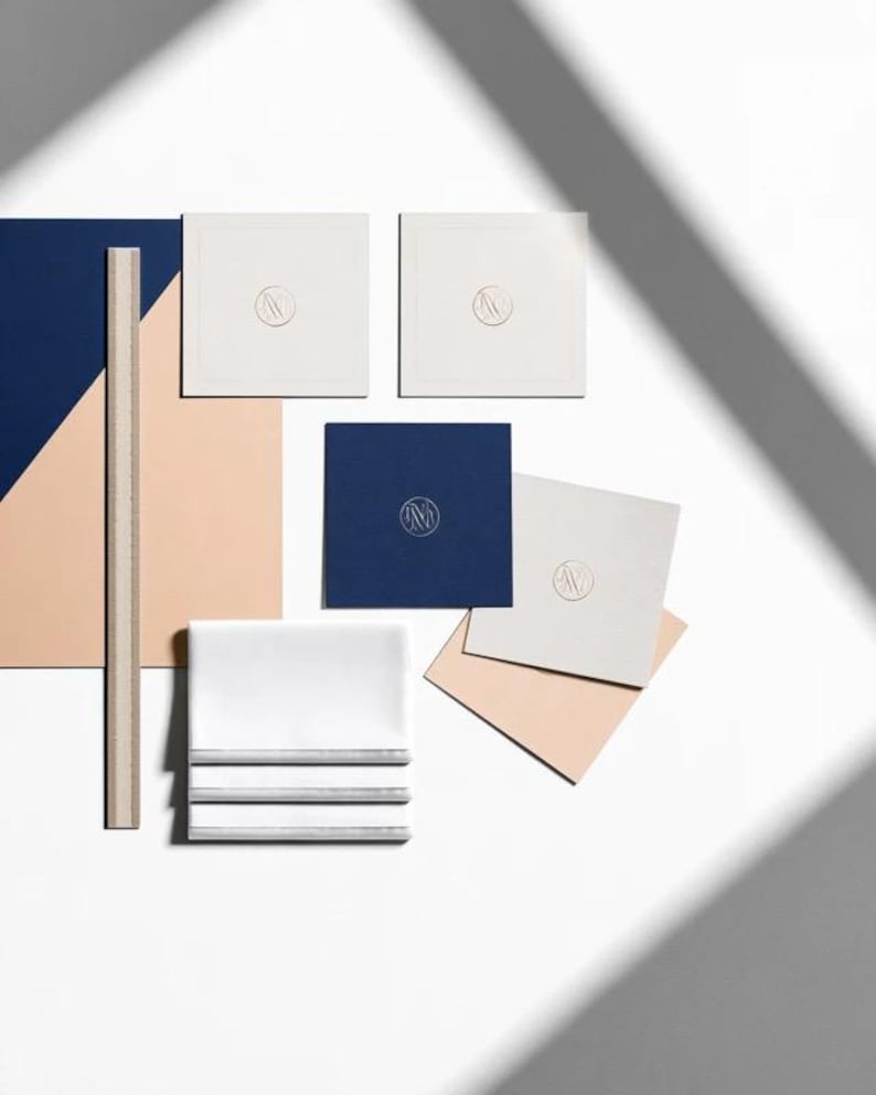 Square navy and cream cocktail napkins with small contrasting monogram letters, displayed with coordinating paper pieces. Digital concept mockup showing a personalised wedding or event table detail.