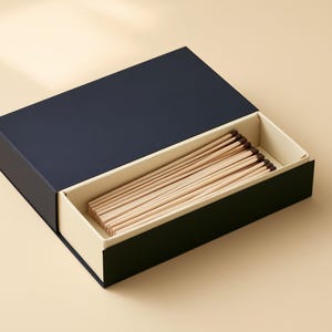 AI-assisted studio design visual of a navy matchbox sleeve with a partly open ivory drawer and wooden matches. Illustrates the 14.5 x 10.5 x 3.5 cm box format; match length and fill are confirmed before ordering.
