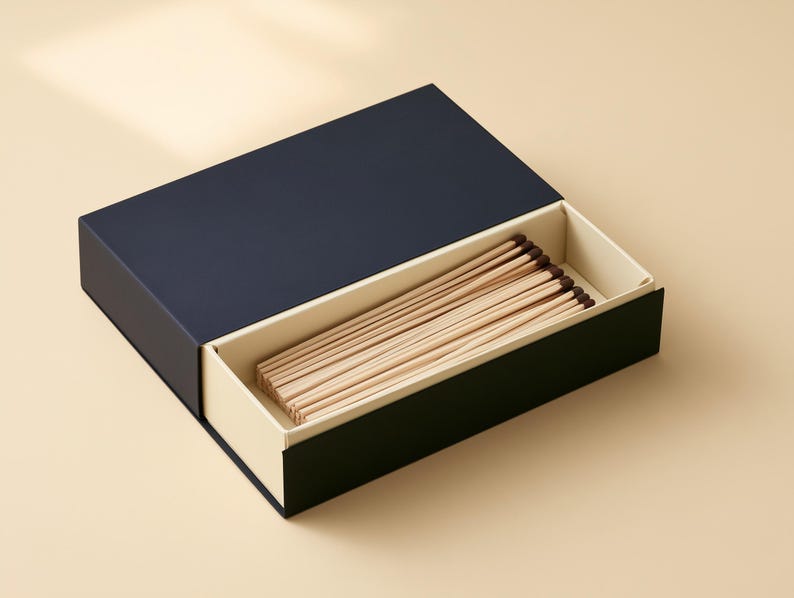 AI-assisted studio design visual of a navy matchbox sleeve with a partly open ivory drawer and wooden matches. Illustrates the 14.5 x 10.5 x 3.5 cm box format; match length and fill are confirmed before ordering.