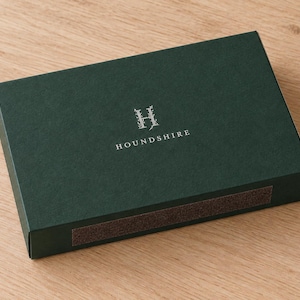 Digital design mockup of one closed forest-green broad drawer matchbox with silver-foil Houndshire monogram on a light oak surface. Optional paid foil finish; illustrative 14.5cm product format, no exposed matches.