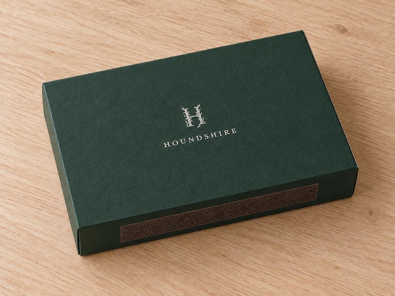 Digital design mockup of one closed forest-green broad drawer matchbox with silver-foil Houndshire monogram on a light oak surface. Optional paid foil finish; illustrative 14.5cm product format, no exposed matches.