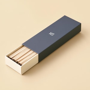 AI-assisted studio design visual of a navy HS example matchbox with an ivory sliding drawer partly open to show wooden matches. Illustrates the 28 x 10.5 x 3 cm outside box format; match length and fill are confirmed before ordering.