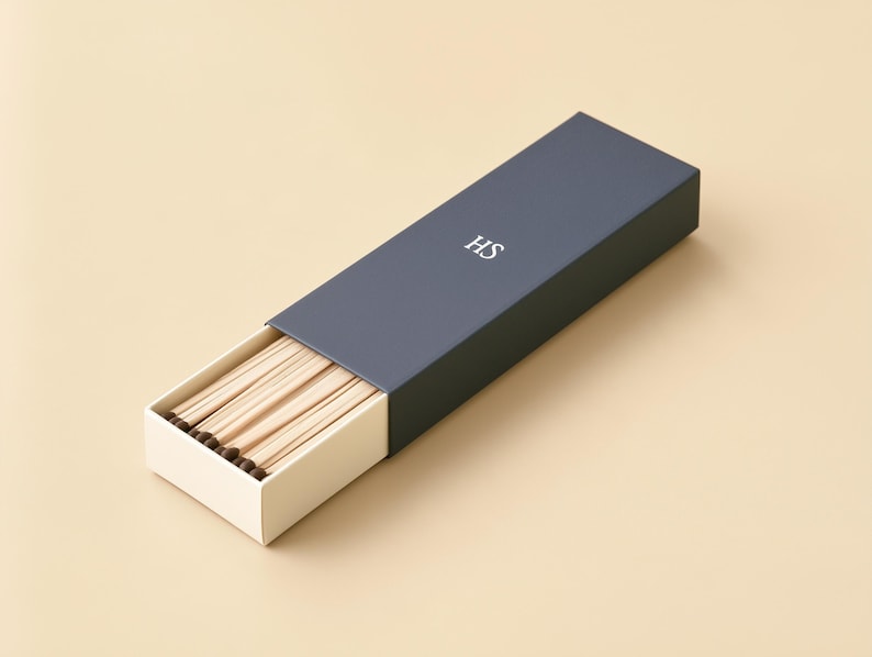 AI-assisted studio design visual of a navy HS example matchbox with an ivory sliding drawer partly open to show wooden matches. Illustrates the 28 x 10.5 x 3 cm outside box format; match length and fill are confirmed before ordering.