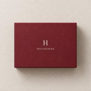 AI-assisted studio illustration of the broad 14.5 x 10.5 x 3.5 cm burgundy matchbox, shown closed from above with an ivory Houndshire sample monogram on the front.
