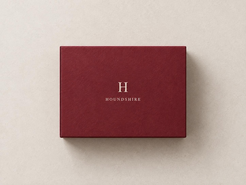 AI-assisted studio illustration of the broad 14.5 x 10.5 x 3.5 cm burgundy matchbox, shown closed from above with an ivory Houndshire sample monogram on the front.