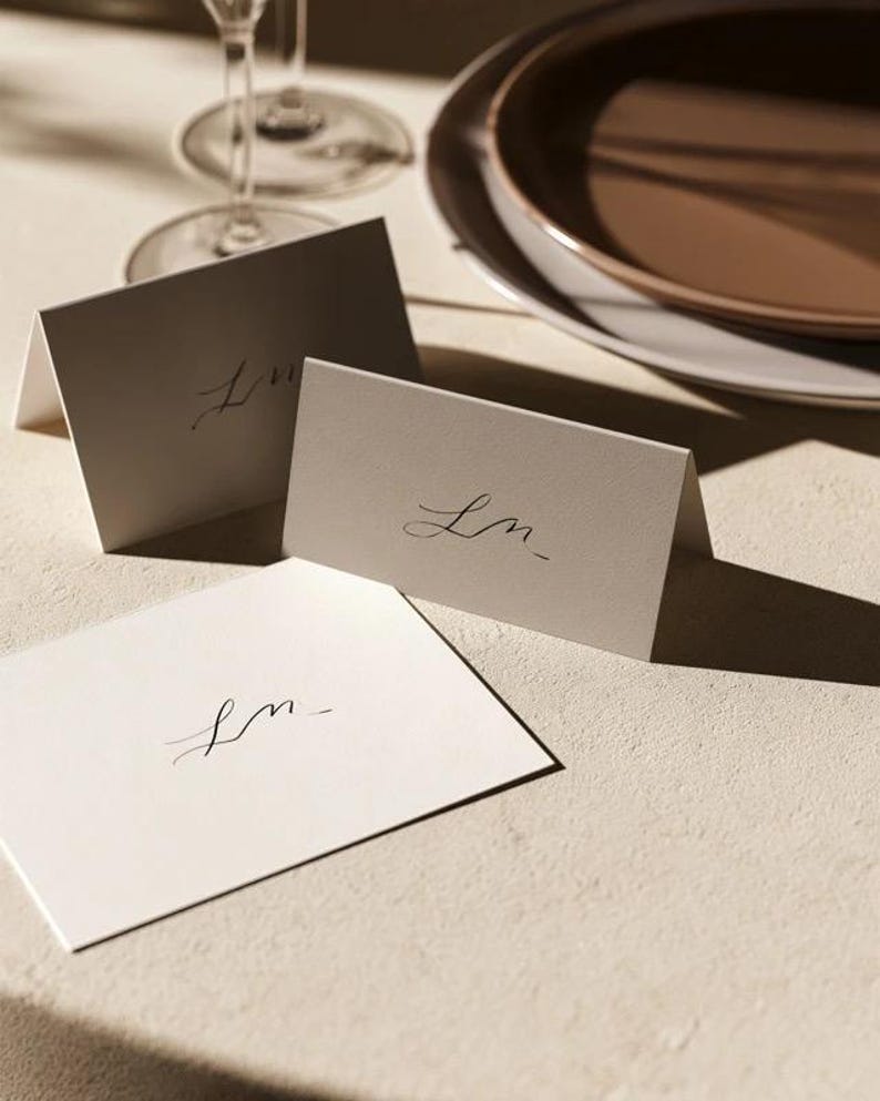 Cream wedding place cards with small dark initials and fine lettering, shown as flat and folded cards on a pale surface. Digital concept mockup of personalised table stationery.