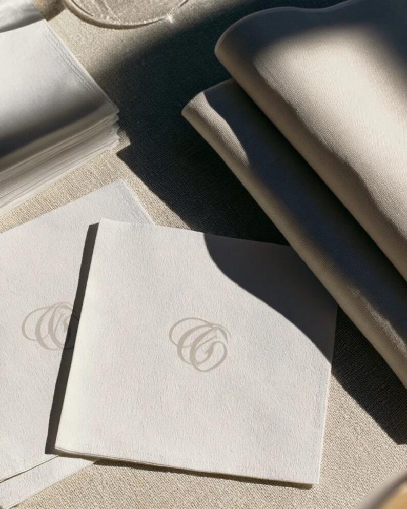 Square navy and cream cocktail napkins with small contrasting monogram letters, displayed with coordinating paper pieces. Digital concept mockup showing a personalised wedding or event table detail.