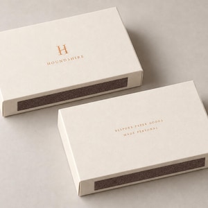 Digital design mockup of two closed ivory broad drawer matchboxes with copper-foil front and reverse branding on a warm-grey studio surface. Optional foil is quoted separately; no exposed matches.