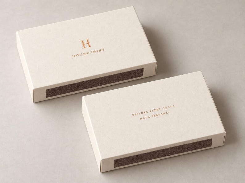 Digital design mockup of two closed ivory broad drawer matchboxes with copper-foil front and reverse branding on a warm-grey studio surface. Optional foil is quoted separately; no exposed matches.