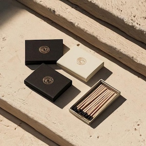 Small rectangular matchbox mockups in a restrained neutral palette, decorated with fine monograms. The styled arrangement shows the paper sleeves and drawer-style construction for personalised event favours.