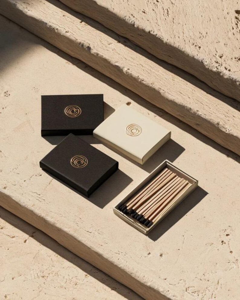 Small rectangular matchbox mockups in a restrained neutral palette, decorated with fine monograms. The styled arrangement shows the paper sleeves and drawer-style construction for personalised event favours.