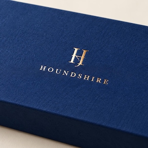 Digital close-up concept of reflective gold-foil Houndshire lettering and monogram on matte navy paper, showing the optional metallic finish and folded matchbox edge. Not a photograph of a manufactured sample.