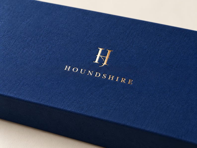 Digital close-up concept of reflective gold-foil Houndshire lettering and monogram on matte navy paper, showing the optional metallic finish and folded matchbox edge. Not a photograph of a manufactured sample.