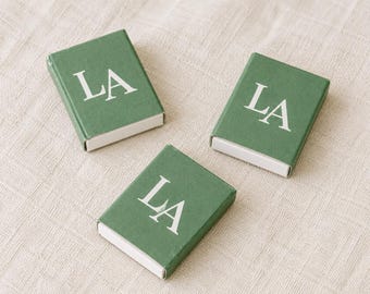 May include: Three rectangular matchboxes with a matte green finish and the white capital letters LA printed on the top surface. Each box features a white inner sliding tray visible at the bottom edge, arranged in a triangular formation.