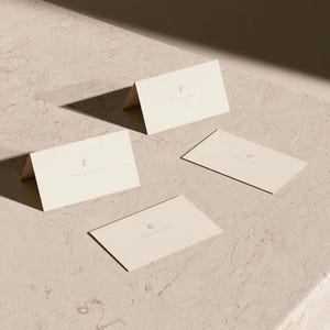 Cream wedding place cards with small dark initials and fine lettering, shown as flat and folded cards on a pale surface. Digital concept mockup of personalised table stationery.