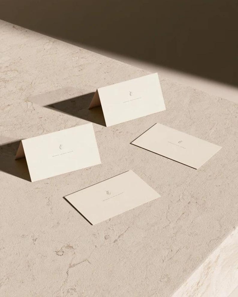 Cream wedding place cards with small dark initials and fine lettering, shown as flat and folded cards on a pale surface. Digital concept mockup of personalised table stationery.