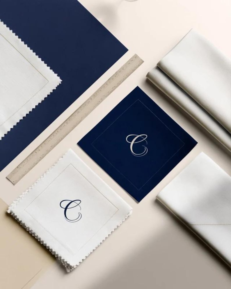 Square navy and cream cocktail napkins with small contrasting monogram letters, displayed with coordinating paper pieces. Digital concept mockup showing a personalised wedding or event table detail.
