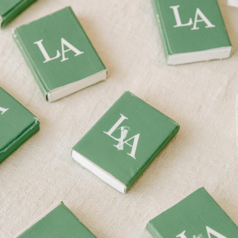 以下が含まれることがあります： 緑色のカバーに白い文字で「LA」とプリントされた長方形のマッチ箱のコレクション。各箱の底面には白い内箱が見えており、一つの箱には文字の間に小さな白いマッチ棒のアイコンが描かれています。