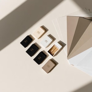 Small rectangular matchbox mockups in a restrained neutral palette, decorated with fine monograms. The styled arrangement shows the paper sleeves and drawer-style construction for personalised event favours.