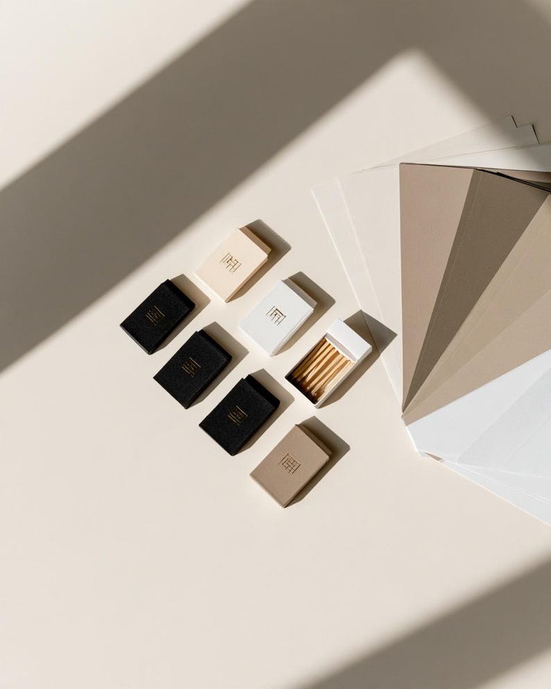 Small rectangular matchbox mockups in a restrained neutral palette, decorated with fine monograms. The styled arrangement shows the paper sleeves and drawer-style construction for personalised event favours.