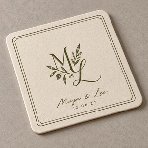 Digital close-up mockup of a square ivory paper coaster with rounded corners, olive ML monogram, fine double-line frame and personalised wording on a plain warm-grey surface, showing the paper texture.