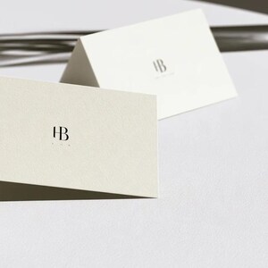Cream wedding place cards with small dark initials and fine lettering, shown as flat and folded cards on a pale surface. Digital concept mockup of personalised table stationery.