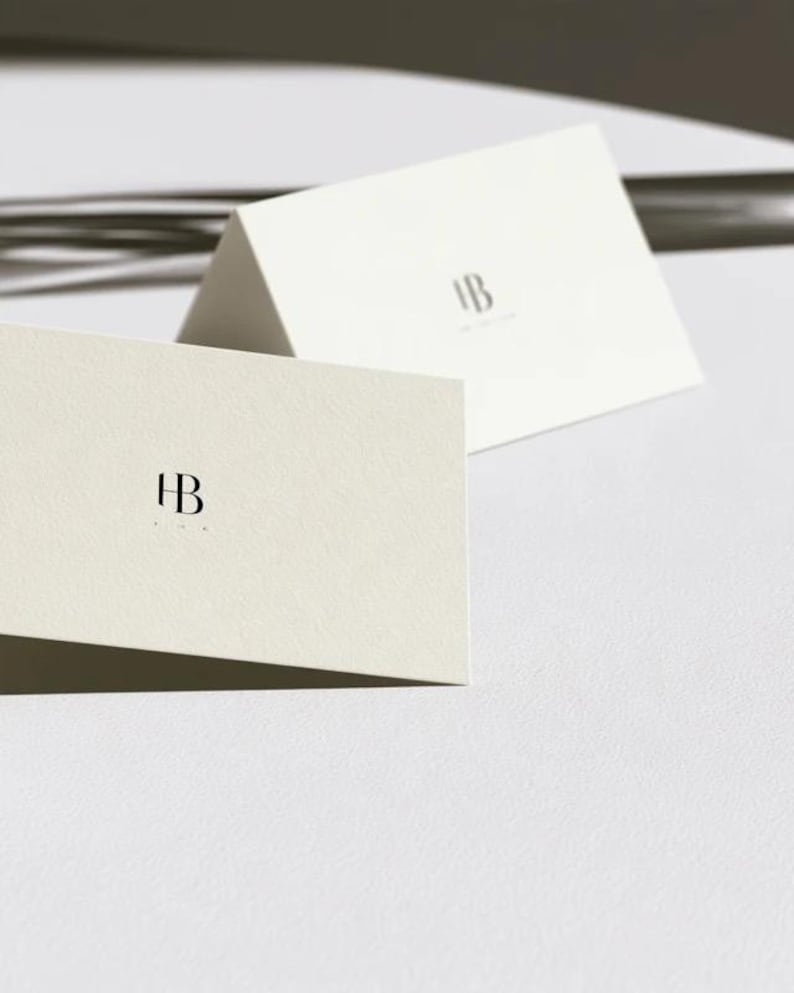 Cream wedding place cards with small dark initials and fine lettering, shown as flat and folded cards on a pale surface. Digital concept mockup of personalised table stationery.